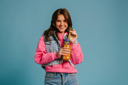 Téléchargez les photos : Joyeux jeune femme en denim porter tenant bouteille avec de la limonade et souriant sur fond bleu - en image libre de droit