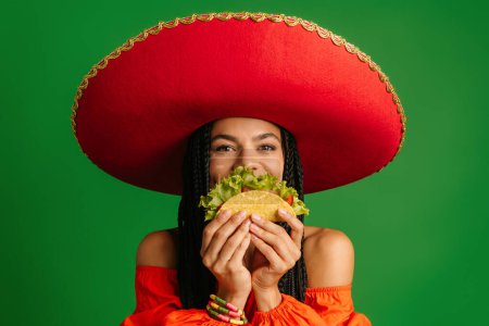 Téléchargez les photos : Femme mexicaine joyeuse dans Sombrero cachant demi-visage derrière taco sur fond vert - en image libre de droit
