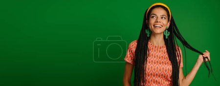 Téléchargez les photos : Jolie jeune femme avec dreadlocs touchant les cheveux et souriant sur fond vert - en image libre de droit