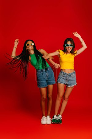 Photo for Full length of two playful young women in colorful wear holding hands while having fun against red background - Royalty Free Image