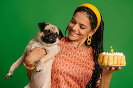 Foto de Atractiva joven hispana que lleva lindo perro pug y pastel de cumpleaños sobre fondo verde - Imagen libre de derechos
