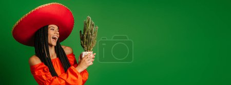 Téléchargez les photos : Belle jeune femme mexicaine dans le Sombrero regardant le cactus et souriant sur fond vert - en image libre de droit