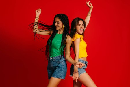 Photo for Two happy young women in colorful wear standing back to back while dancing against red background - Royalty Free Image