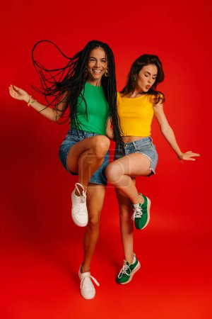 Photo for Full length of two happy young women in colorful wear dancing against red background - Royalty Free Image