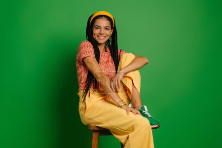 Photo for Beautiful young woman with dreadlocs looking at camera while sitting on the chair against green background - Royalty Free Image