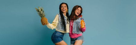 Téléchargez les photos : Deux jeunes femmes ludiques tenant ananas et bouteille de limonade sur fond bleu - en image libre de droit