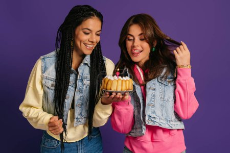 Foto de Dos hermosas mujeres jóvenes mirando el pastel de cumpleaños y sonriendo sobre el fondo púrpura - Imagen libre de derechos