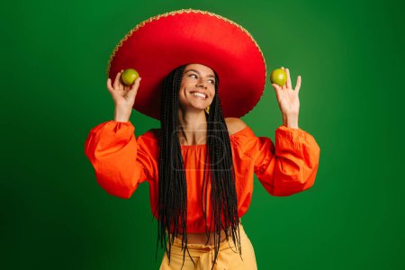 Foto de Joven mexicana alegre en Sombrero sosteniendo limas frescas y sonriendo sobre fondo verde - Imagen libre de derechos