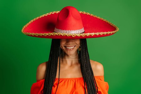 Téléchargez les photos : Superbe jeune femme mexicaine couvrant demi-visage avec Sombrero et souriant sur fond vert - en image libre de droit