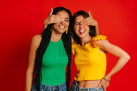 Photo for Two joyful young women in colorful wear covering eyes to each other and smiling against red background - Royalty Free Image