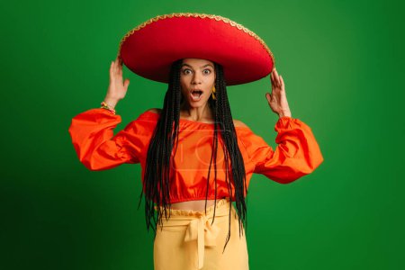 Foto de Sorprendida joven mexicana en Sombrero mirando a la cámara mientras está de pie sobre fondo verde - Imagen libre de derechos