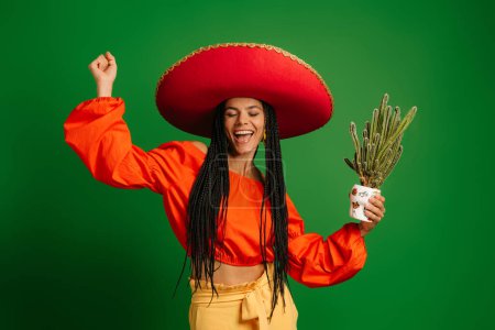 Téléchargez les photos : Belle femme mexicaine dans le Sombrero tenant des cactus et dansant sur fond vert - en image libre de droit