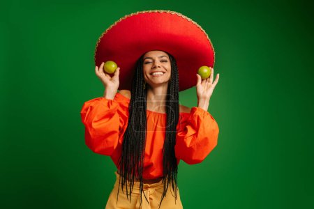 Foto de Atractiva joven mexicana en Sombrero sosteniendo limas frescas y sonriendo sobre fondo verde - Imagen libre de derechos