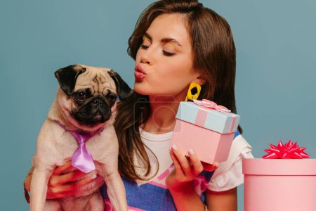 Téléchargez les photos : Belle jeune femme caressant chien mignon et tenant boîte cadeau tout en étant assis au bureau rose sur fond bleu - en image libre de droit