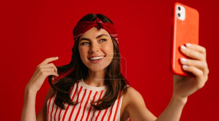 Téléchargez les photos : Belle jeune femme hipster faisant selfie par téléphone mart et souriant tout en se tenant debout sur fond rouge - en image libre de droit