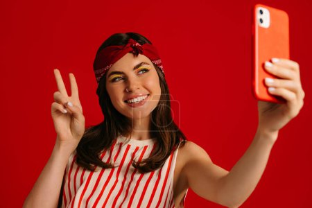Téléchargez les photos : Joyeuse jeune femme hipster faisant selfie par téléphone mart et souriant tout en se tenant debout sur fond rouge - en image libre de droit