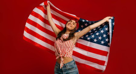Téléchargez les photos : Joyeuse jeune femme hipster portant le drapeau américain tout en se tenant sur fond rouge - en image libre de droit