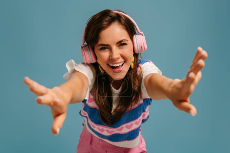 Téléchargez les photos : Belle jeune femme dans les écouteurs profitant de la musique et souriant tout en se tenant debout sur fond bleu - en image libre de droit