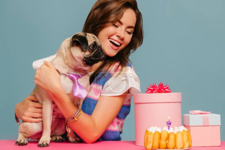 Photo for Joyful woman petting cute dog and looking at the birthday cake while sitting at the pink desk on blue background - Royalty Free Image