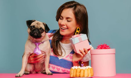 Foto de Feliz joven mujer abrazando lindo perro y sosteniendo caja de regalo mientras está sentado en el escritorio rosa sobre fondo azul - Imagen libre de derechos