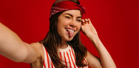 Foto de Juguetona joven hipster mujer haciendo selfie mientras mantiene el brazo extendido sobre fondo rojo - Imagen libre de derechos