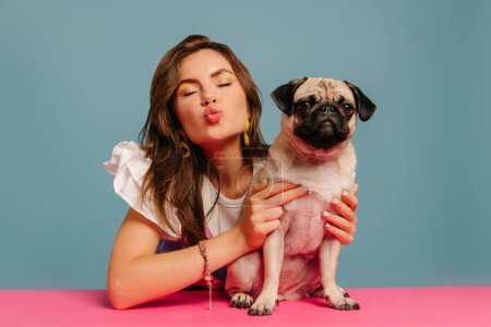 Téléchargez les photos : Belle femme soufflant comme baiser et étreignant chien chiot mignon tout en étant assis au bureau rose sur fond bleu - en image libre de droit