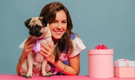 Foto de Atractiva mujer joven acariciando perro lindo pug y sonriendo mientras está sentado en el escritorio rosa sobre fondo azul - Imagen libre de derechos
