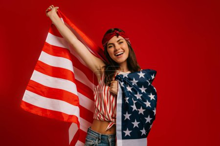 Téléchargez les photos : Jolie jeune femme hipster portant le drapeau américain et souriant tout en se tenant debout sur fond rouge - en image libre de droit