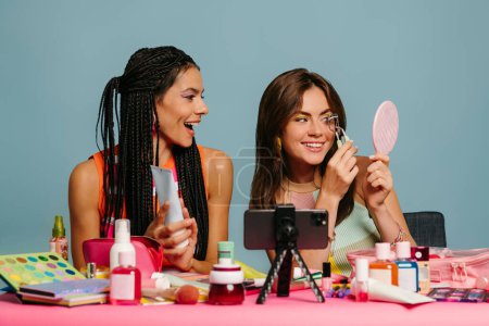 Foto de Dos alegres influencers femeninas transmitiendo un vlog de belleza mientras están sentadas en el escritorio contra el fondo azul - Imagen libre de derechos