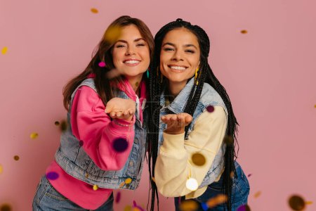 Foto de Dos mujeres jóvenes alegres soplando confeti colorido y sonriendo sobre fondo rosa - Imagen libre de derechos