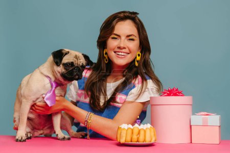 Téléchargez les photos : Joyeuse femme caressant chien mignon et célébrer l'anniversaire tout en étant assis au bureau rose sur fond bleu - en image libre de droit