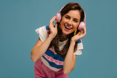 Téléchargez les photos : Joyeux jeune femme dans les écouteurs jouissant de la musique tout en se tenant sur fond bleu - en image libre de droit