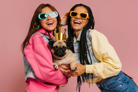 Foto de Dos mujeres jóvenes felices en gafas de moda que llevan lindo perro pug en corona divertida contra el fondo rosa - Imagen libre de derechos