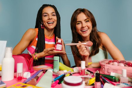 Foto de Dos presentadoras femeninas felices mostrando varios productos de belleza mientras están sentadas en el escritorio sobre fondo azul - Imagen libre de derechos