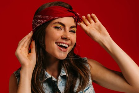 Téléchargez les photos : Attrayant jeune femme hipster avec maquillage coloré ajustement bandana et sourire sur fond rouge - en image libre de droit