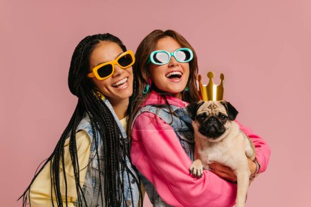 Téléchargez les photos : Deux jeunes femmes à la mode dans des lunettes à la mode portant chien carlin mignon et souriant sur fond rose - en image libre de droit