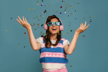 Téléchargez les photos : Joyeuse jeune femme dans des écouteurs et des lunettes à la mode profitant de la musique tout en dansant sur fond bleu - en image libre de droit