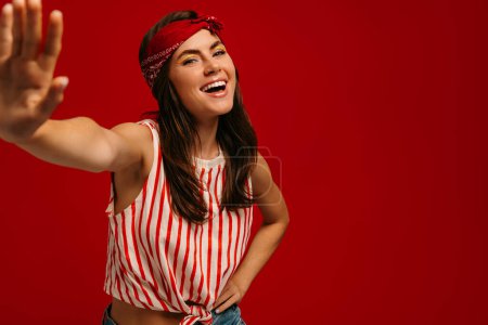 Téléchargez les photos : Joyeux jeune femme hipster étirant la main et souriant tout en se tenant debout sur fond rouge - en image libre de droit
