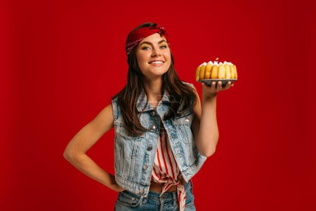 Foto de Feliz joven mujer sosteniendo pastel de cumpleaños y mirando a la cámara con sonrisa en el fondo rojo - Imagen libre de derechos