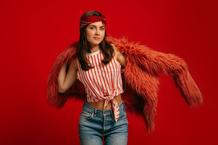 Photo for Fashionable young hipster woman wearing fluffy coat and smiling while standing on red background - Royalty Free Image
