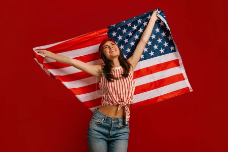 Téléchargez les photos : Joyeux jeune femme hipster portant le drapeau américain tout en se tenant sur fond rouge - en image libre de droit