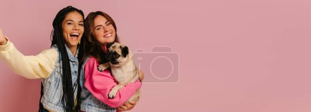 Téléchargez les photos : Deux jeunes femmes joyeux portant chien chiot mignon et faire selfie sur fond rose - en image libre de droit