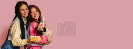 Téléchargez les photos : Deux jeunes femmes joyeux collant tout en portant chien chiot mignon et gâteau d'anniversaire sur fond rose - en image libre de droit