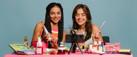 Foto de Dos influencers femeninas felices examinando productos de belleza mientras que transmiten en línea contra fondo azul - Imagen libre de derechos