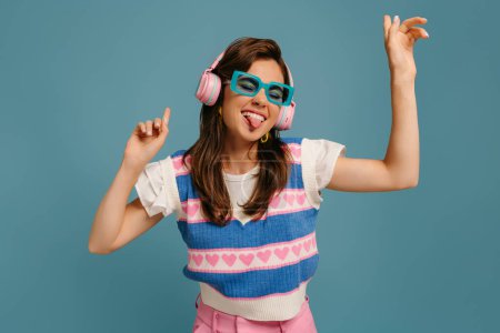 Foto de Jovencita juguetona con auriculares y gafas de moda disfrutando de la música mientras baila sobre fondo azul - Imagen libre de derechos