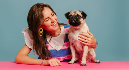 Téléchargez les photos : Attrayant jeune femme étreignant chien chiot mignon et souriant tout en étant assis au bureau rose sur fond bleu - en image libre de droit