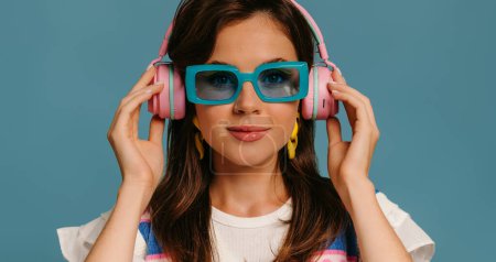 Téléchargez les photos : Portrait de belle jeune femme dans des écouteurs et des lunettes à la mode profitant de la musique sur fond bleu - en image libre de droit