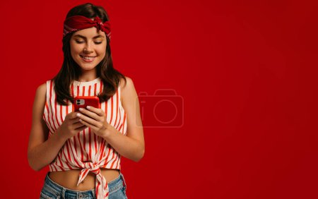 Foto de Elegante joven hipster mujer mensaje de texto en el teléfono inteligente y sonriendo mientras está de pie sobre fondo rojo - Imagen libre de derechos