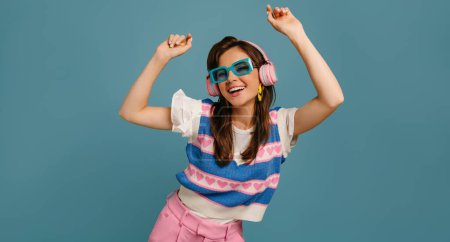 Foto de Joven alegre con auriculares y gafas de moda disfrutando de la música mientras baila sobre fondo azul - Imagen libre de derechos