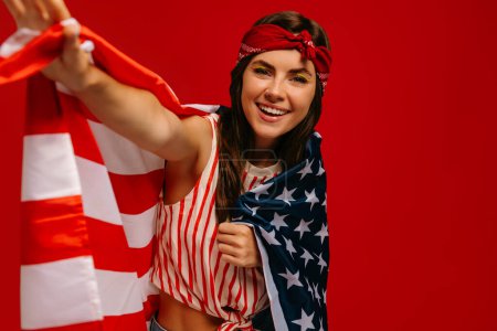 Foto de Hermosa joven hipster mujer llevando bandera americana y sonriendo mientras está de pie sobre fondo rojo - Imagen libre de derechos
